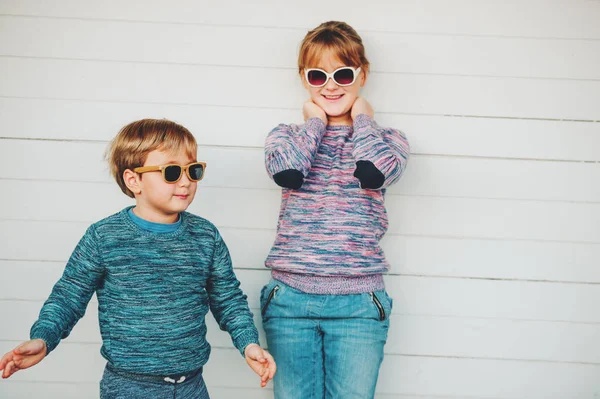 Grupo de dos niños divertidos jugando juntos al aire libre, niño y niña posando sobre fondo de madera blanca, hermano y hermana vistiendo pullovers a juego, moda para niños, prendas de punto . — Foto de Stock