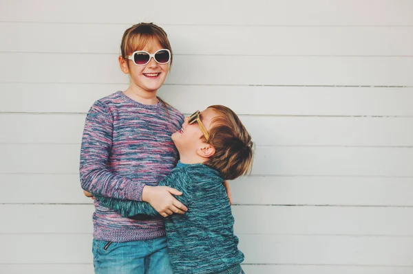 Groep van twee grappige kinderen samen buiten spelen, kleine jongen en meisje poseren tegen witte houten achtergrond, broer en zus dragen bijpassende truien, mode voor kinderen, breigoed. — Stockfoto