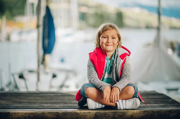 Außenporträt des süßen kleinen 6-jährigen Jungen, der an einem schönen sonnigen Tag in einem Hafen ruht und rote Körperwärmer trägt — Stockfoto