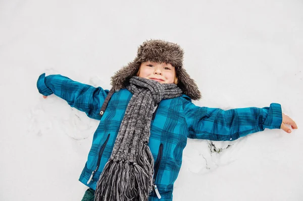 Söt liten pojke spelar i Vinterparken. Kid har roligt utomhus, sitter på snön, bär varma blå jacka, mössa och halsduk — Stockfoto