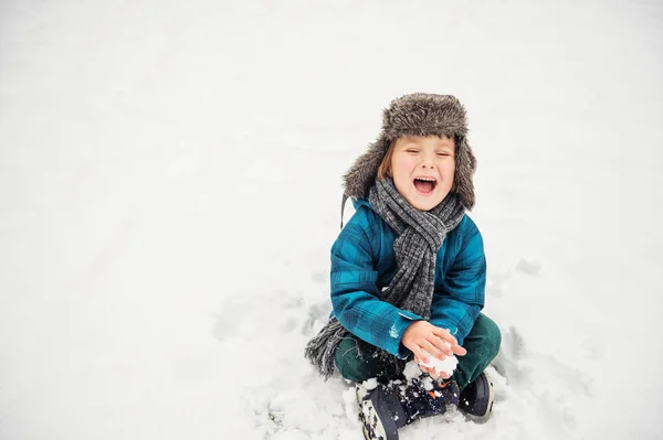 可爱的小男孩在冬季公园玩。孩子在户外玩得开心，坐在雪地上，穿着温暖的蓝色外套、 帽子和围巾 — 图库照片