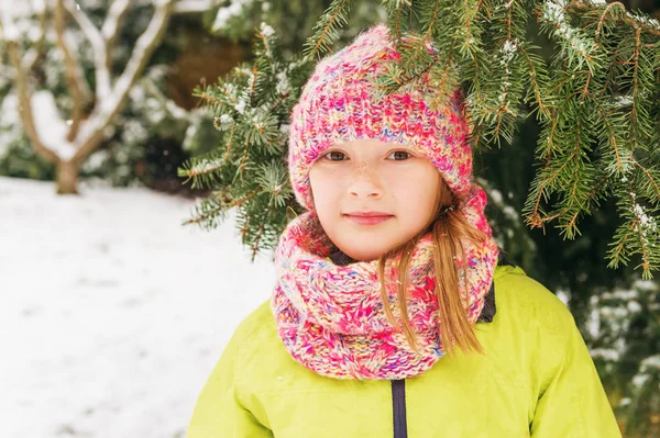 Yeşil ceket, renkli örgü şapka ve atkı giyen sevimli küçük bir kız kar yağışı altında kış portresi — Stok fotoğraf