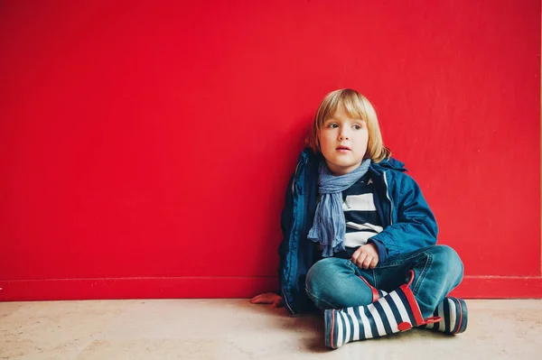 Retrato de moda de adorable niño de 4-5 años, con chaqueta azul, bufanda y botas de lluvia rayas, sentado en el suelo contra la pared de color rojo brillante — Foto de Stock