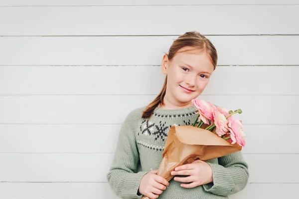 Ritratto di adorabile bambina di 8-9 anni, vestita con un caldo pullover verde, con in mano fiori rosa primaverili, in piedi su uno sfondo di legno bianco, concetto di festa della mamma — Foto Stock