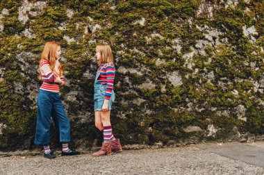 Outdoor portrait of two funny preteen girls wearing denim shorts, stripe roll neck pullovers and long socks, posing against moss wall, fashion for teens clipart
