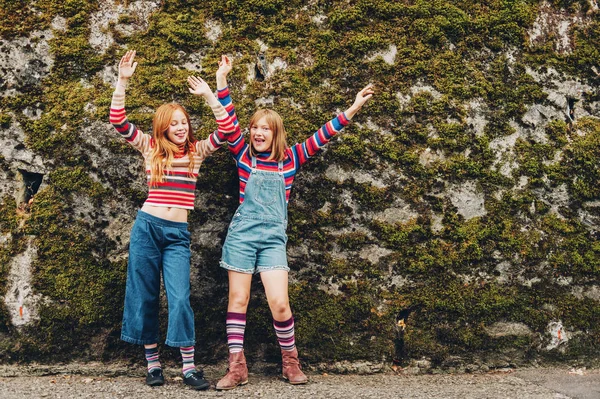 Retrato ao ar livre de duas meninas pré-adolescentes engraçadas usando shorts de ganga, pulôveres de pescoço de rolo de listra e meias longas, posando contra a parede de musgo, moda para adolescentes — Fotografia de Stock