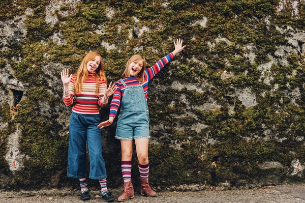 Retrato ao ar livre de duas meninas pré-adolescentes engraçadas usando shorts de ganga, pulôveres de pescoço de rolo de listra e meias longas, posando contra a parede de musgo, moda para adolescentes — Fotografia de Stock