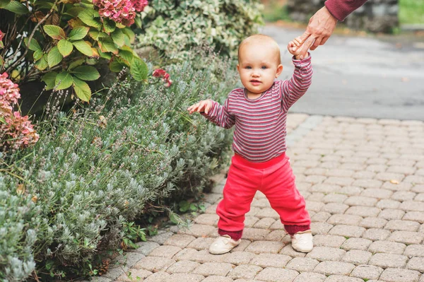 Bedårande flicka 9-12 månader gamla spelar utanför, klädd i rosa kropp och joggare, hålla mammas hand. Barnets första steg, kid lära sig gå — Stockfoto