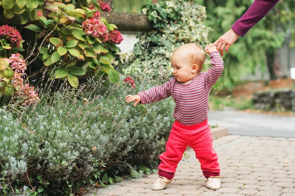 Niña adorable de 9-12 meses jugando al aire libre, vistiendo cuerpo rosa y corredores, sosteniendo la mano de la madre. Primeros pasos del niño, niño aprendiendo a caminar — Foto de Stock