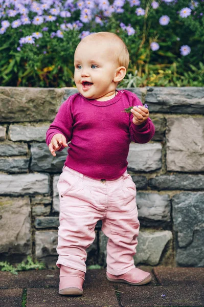 Outdoor Portret van schattige 9-12 maand oude babymeisje spelen met paarse bloemen, het dragen van roze broek, leren laarzen en paarse body — Stockfoto