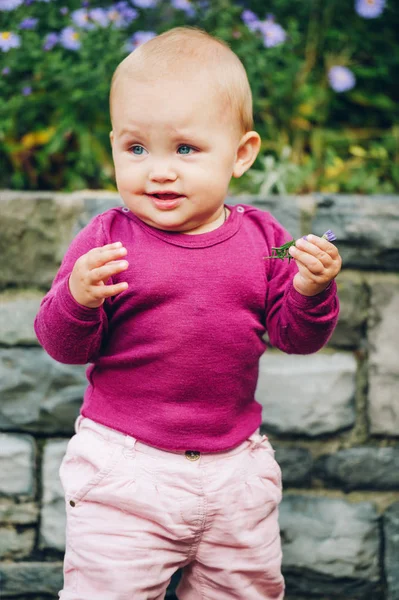 Retrato al aire libre de la adorable niña de 9-12 meses jugando con flores moradas, usando pantalones rosados y cuerpo morado —  Fotos de Stock