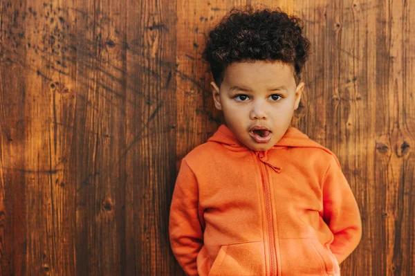 Retrato ao ar livre de menino adorável criança posando fora contra o fundo de madeira marrom, vestindo jaqueta com capuz laranja — Fotografia de Stock