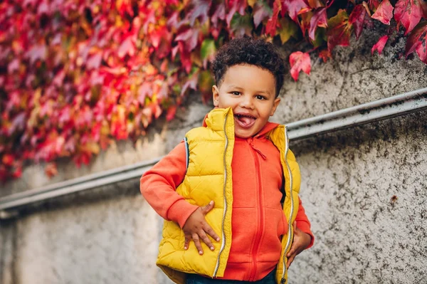 Portrait d'adorable jeune garçon africain jouant à l'extérieur par une belle journée d'automne, portant une veste à capuche orange vif et une veste gilet jaune — Photo