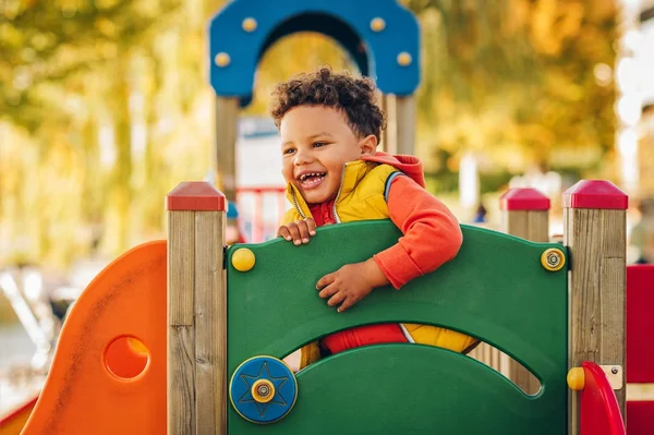 Очаровательный маленький 1-2-летний мальчик развлекается на детской площадке, ребенок в оранжевой куртке с капюшоном и желтом жилете — стоковое фото
