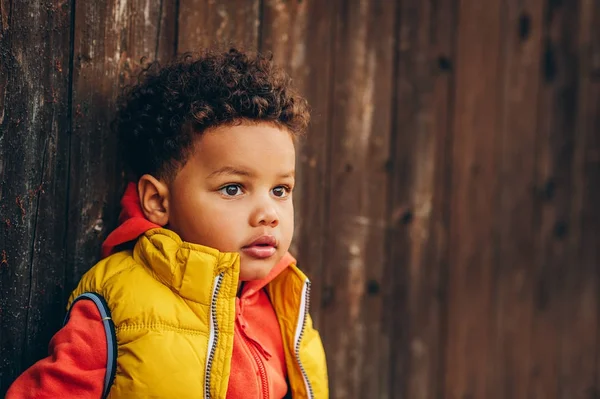Retrato ao ar livre de menino adorável criança posando fora contra fundo de madeira marrom, vestindo jaqueta com capuz laranja qualquer casaco de colete amarelo brilhante — Fotografia de Stock