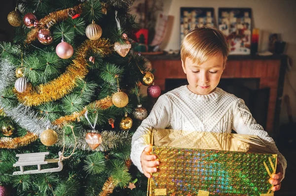 Feche o retrato do menino feliz jogando ao lado da árvore de Natal, vestindo pulôver quente branco, segurando grande presente de pacote dourado — Fotografia de Stock