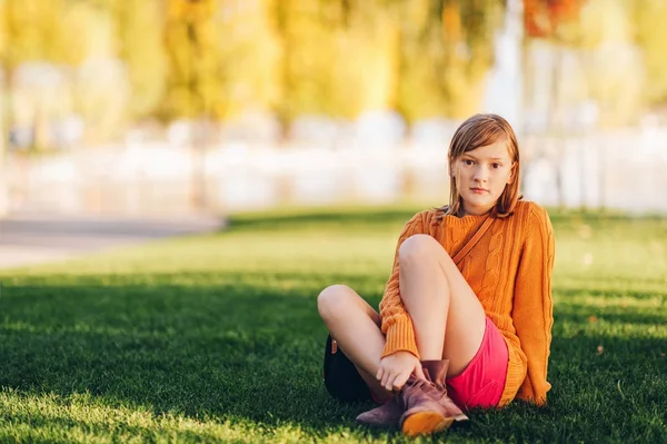 Outdoor portrait of sweet little preteen kid girl, sitting on bright green lawn, wearing orange pullover and red shorts — Stock Photo, Image