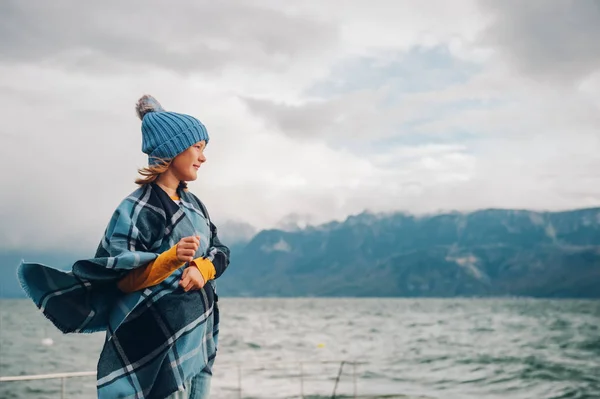 Süßes kleines Mädchen, das an einem sehr windigen Tag am See spielt, mit blauem Hut und kariertem Poncho. Aufnahme vom Genfer See, Lausanne, Schweiz — Stockfoto