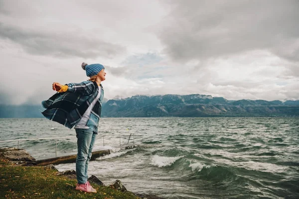 Douce petite fille jouant au bord du lac par une journée très venteuse, portant un chapeau bleu et poncho à carreaux. Photo prise sur le lac Léman, Lausanne, Suisse — Photo