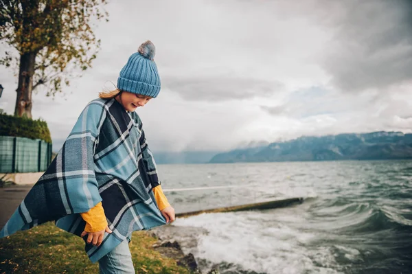 Γλυκό κοριτσάκι παίζει από τη λίμνη σε μια πολύ θυελλώδη ημέρα, φορώντας μπλε καπέλο και καρό poncho. Εικόνα που λαμβάνεται στη λίμνη Γενεύη, Λωζάνη, Ελβετία — Φωτογραφία Αρχείου