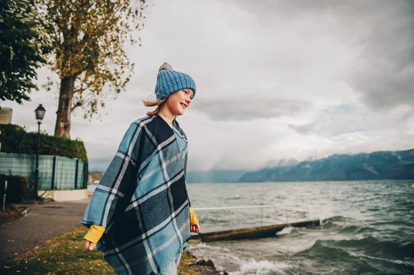 Söt liten flicka spelar vid sjön på en mycket blåsig dag, klädd i blå hatt och pläd poncho. Bild tagen vid Genèvesjön, Lausanne, Schweiz — Stockfoto