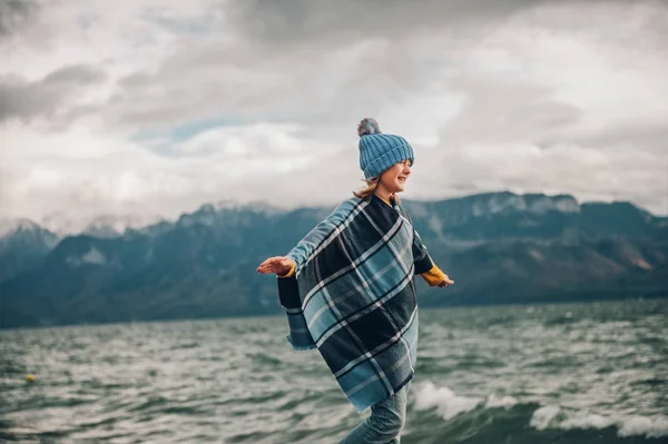 Söt liten flicka spelar vid sjön på en mycket blåsig dag, klädd i blå hatt och pläd poncho. Bild tagen vid Genèvesjön, Lausanne, Schweiz — Stockfoto