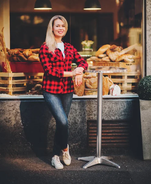 Retrato al aire libre de una hermosa mujer rubia con camisa a cuadros roja, posando al lado de la panadería — Foto de Stock