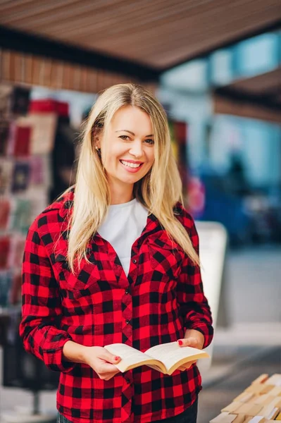 Feliz joven rubia leyendo mujer en una librería, vistiendo camisa a cuadros roja — Foto de Stock