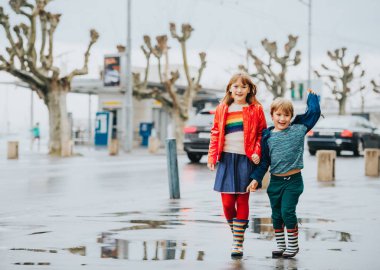 İki komik çocuğu, küçük kardeşim ve kız kardeşi birlikte erken ilkbaharda yağmur altında bir şehirde oynamak. Yağmur ceket ve çizmeler giyen çocuk