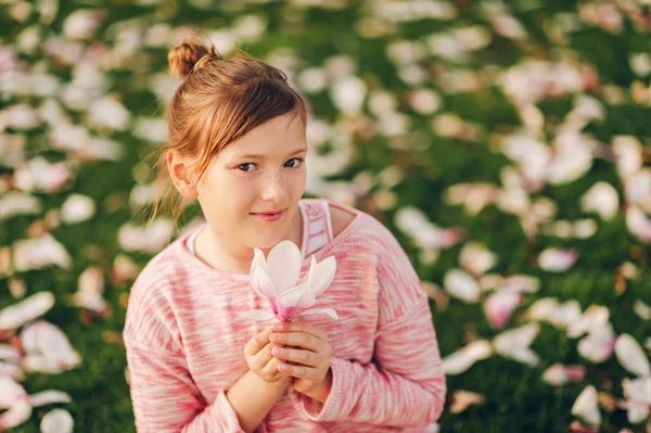 春の花の花びらと緑の草の上に座って 美しいモクレンの花を保持している ピンクのトレーナー 子供のためのファッションを着ているかわいい女の子の肖像画 — ストック写真