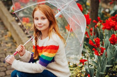 Gökkuşağı kazak, bir yağmurlu günde, moda kız dışında oynayan çocuk giyen şeffaf şemsiye tutan çok güzel kızıl saçlı çocuk kız portresi açık bahar