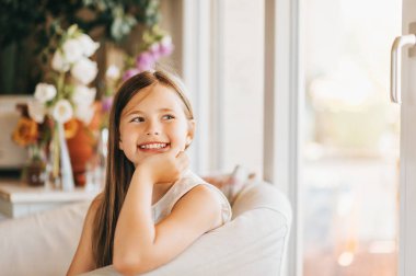 Sevimli küçük kız evde pencere yanında beyaz bir sandalyede oturan