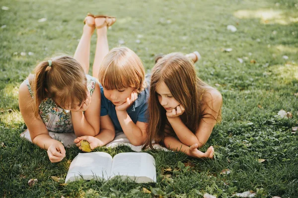 集团的三个孩子躺在绿色的草地上 一起看故事书 儿童教育 — 图库照片