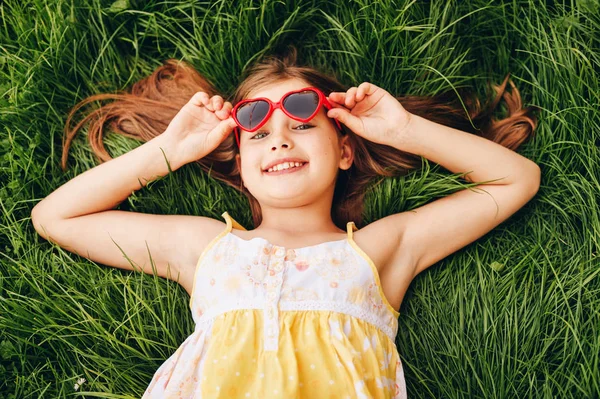 屋外のポートレート中心を身に着けているかわいい女の子の形のサングラス 新鮮な緑の草の上に横たわる 子供たち 夏のお楽しみ 幸せな子供時代のためのパーティー トップ ビュー — ストック写真