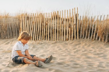 Komik çocuk çocuk deniz kum plajı üzerinde günbatımında dinleniyor. Polo, deni, şort ve sandalet ayakkabı giyen genç çocuk Güney Fransa, yaz tatilinin tadını çıkarıyor