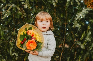 Noel çiçeği taşıyan küçük şirin bir çocuğun portresi, Noel ağacının yanında duruyor.