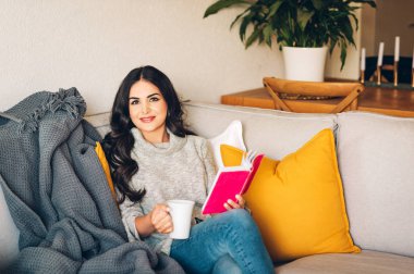 Genç, güzel bir kadın bir kanepede dinleniyor, elinde çay ya da kahve tutuyor, kitap okuyor.