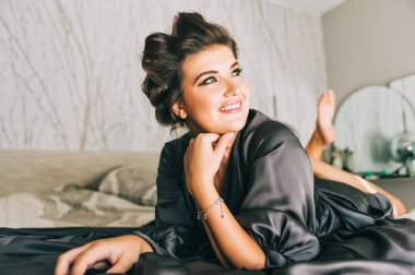 Mutlu genç bir kadının portresi yatak odasında dinleniyor, gri bir gecelik giyiyor, kafasında bigudiler var.