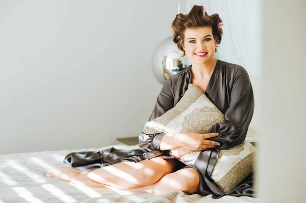 Interior portrait of happy young woman relaxing in bedroom, wearing grey nightie, curlers on the head, holding a pillow