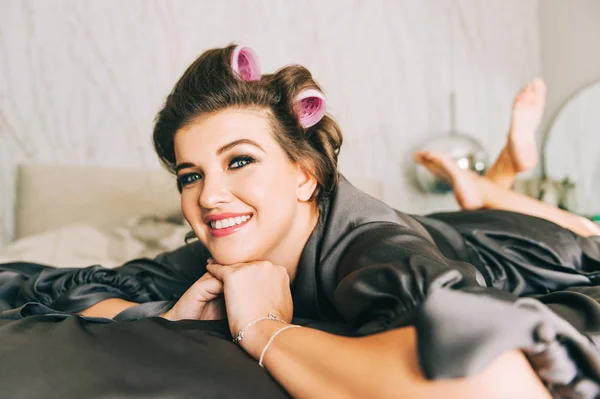 Interior portrait of happy young woman relaxing in bedroom, wearing grey nightie, curlers on the head