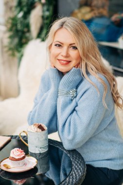 Güzel bir kadının açık hava portresi. Terasta rahat bir kafe ya da çay odasında, elinde bir fincan sıcak çikolata, mavi örgü kazak giyiyor.