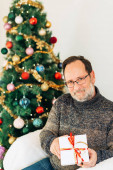Portrét muže středního věku pózujícího vedle vánočního stromku, v teplém svetru a brýlích, držícího dárek v bílé krabici s červenou stuhou 