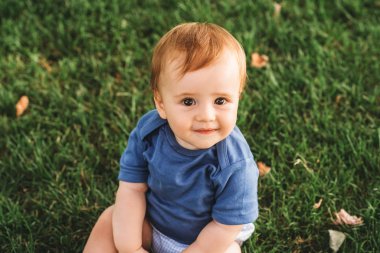 Açık hava portresi, yaz parkında oynayan, yeşil çimlerde oturan, mavi giysili, sevimli kızıl saçlı bebek.