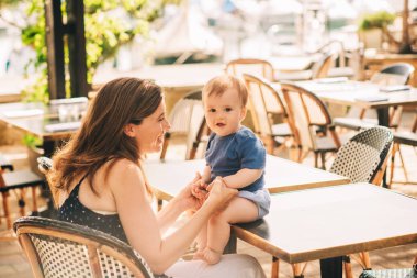 Genç, güzel bir anne, yaz kafeteryasında bebekle oynuyor, çocuk masada oturuyor, annesinin elini tutuyor.
