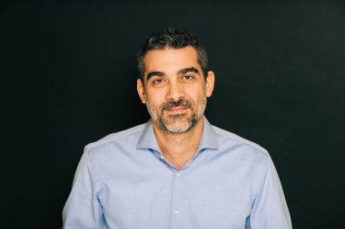 Resmi mavi gömlek giyen, siyah arka planda poz veren yakışıklı bir adamın stüdyo portresi.