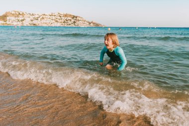 Deniz kenarında oynayan komik küçük çocuk, güneş kremi sürmüş yelek giyiyor, çocuklarla aile tatili yapıyor.