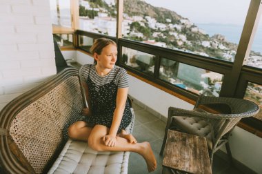 Genç kız kanepede dinleniyor ve balkondan güzel manzaraya hayran, deniz kenarında yaz tatili, çocuklarla aile seyahati.