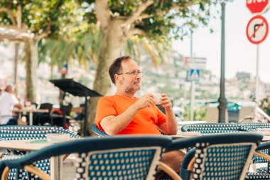 Orta yaşlı, mutlu bir adam. Yazlık kafede dinleniyor. Elinde bir fincan kahve, üzerinde turuncu tişört ve gözlük var.