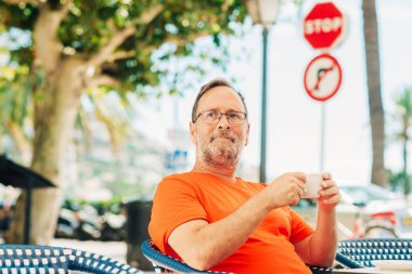 Orta yaşlı, mutlu bir adam. Yazlık kafede dinleniyor. Elinde bir fincan kahve, üzerinde turuncu tişört ve gözlük var.