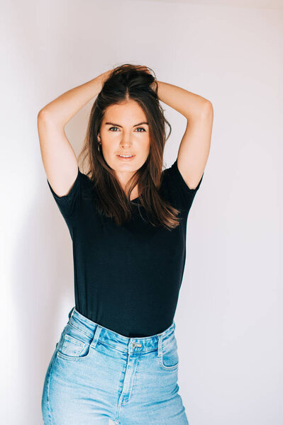 Indoor portrait of beautiful young woman, wearing black t-shirt and high waist jeans, posing on white background, holding hands on head